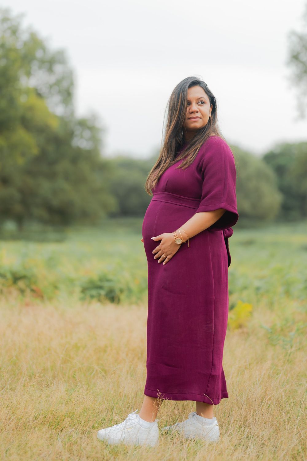 Pregnant woman in burgundy dress standing in a green field, cradling her baby bump outdoors.