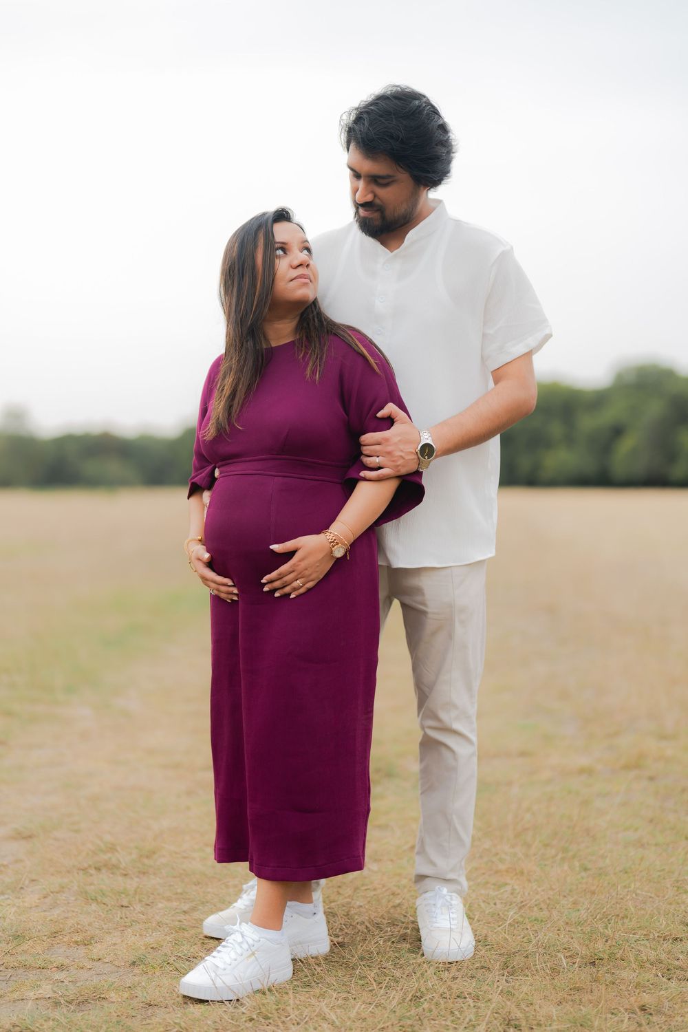 Pregnant woman in purple dress with partner outdoors in a field, sharing a tender moment during maternity photoshoot.
