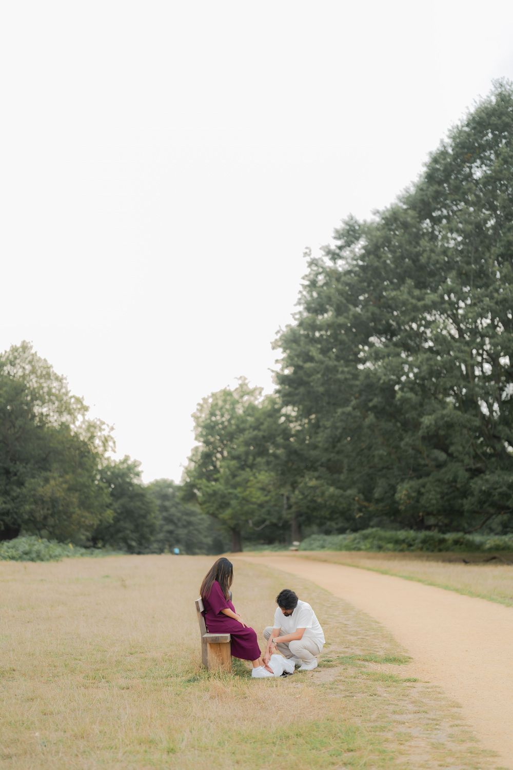 Couple sitting together on a bench along a winding path surrounded by lush green trees in a serene park setting.