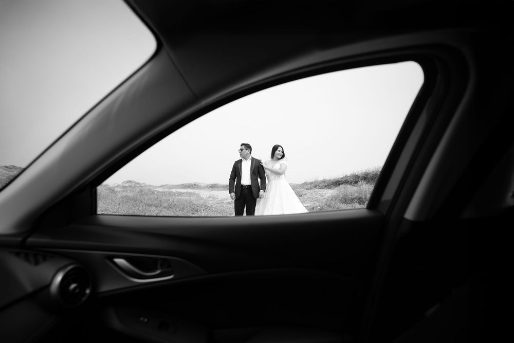 Black and white photo of bride and groom holding hands outdoors, framed through a car window.
