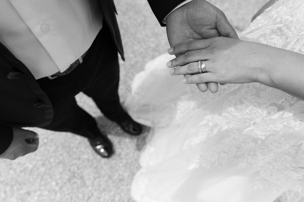 Black and white wedding photo of bride and groom holding hands, showcasing wedding rings on lace bridal gown.