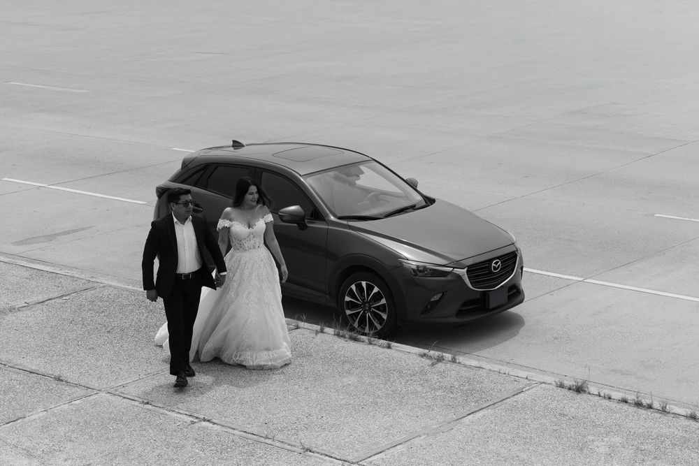 Black and white photo of a bride and groom walking hand in hand beside a Mazda CX-3 on their wedding day.