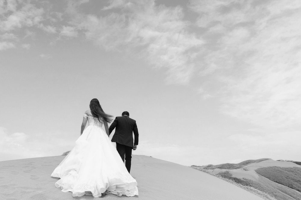 Black and white photo of a bride and groom holding hands walking across sand dunes on their wedding day.