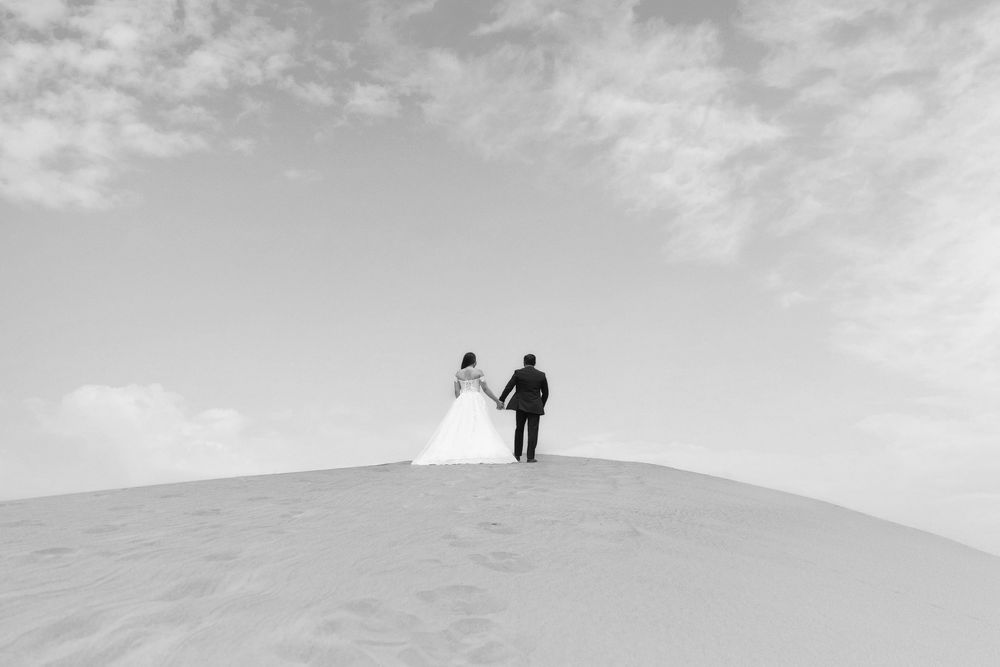 Black and white photo of a bride and groom holding hands atop a sand dune against a cloudy sky.