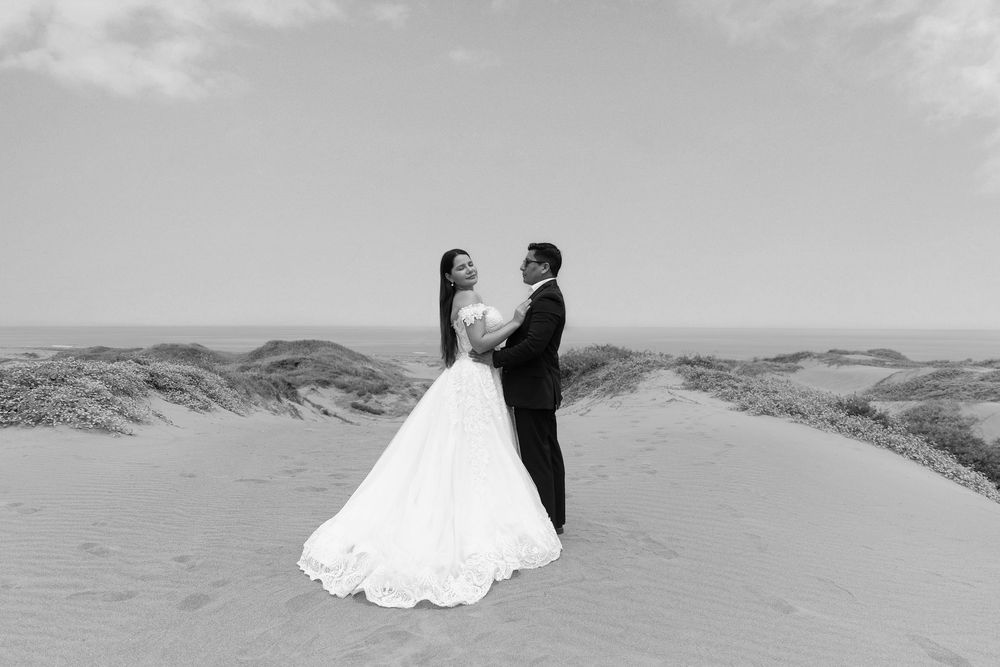 Black and white photo of a bride and groom embracing on sand dunes, bride in flowing gown holding bouquet.