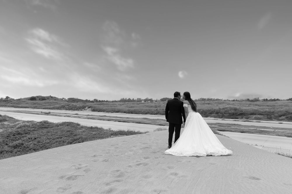 Black and white photo of a bride and groom walking hand in hand along a sandy path with open fields in the background.