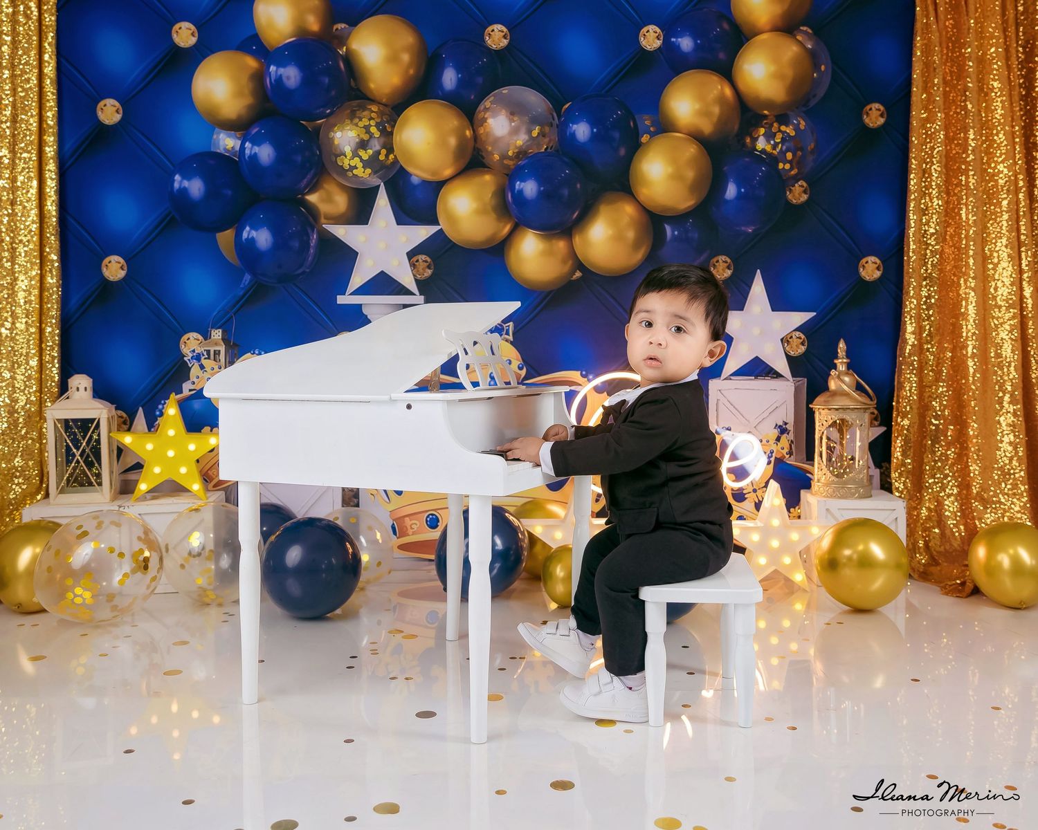 Toddler boy in black suit plays white grand piano surrounded by blue and gold balloons and stars.