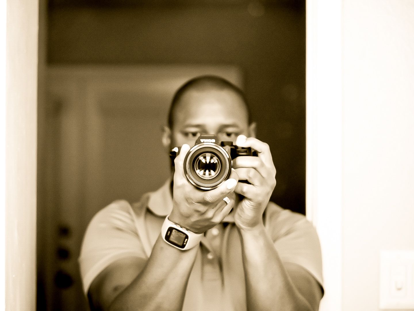 Man taking a mirror selfie with a vintage film camera, wearing a watch, in a sepia-toned photograph.