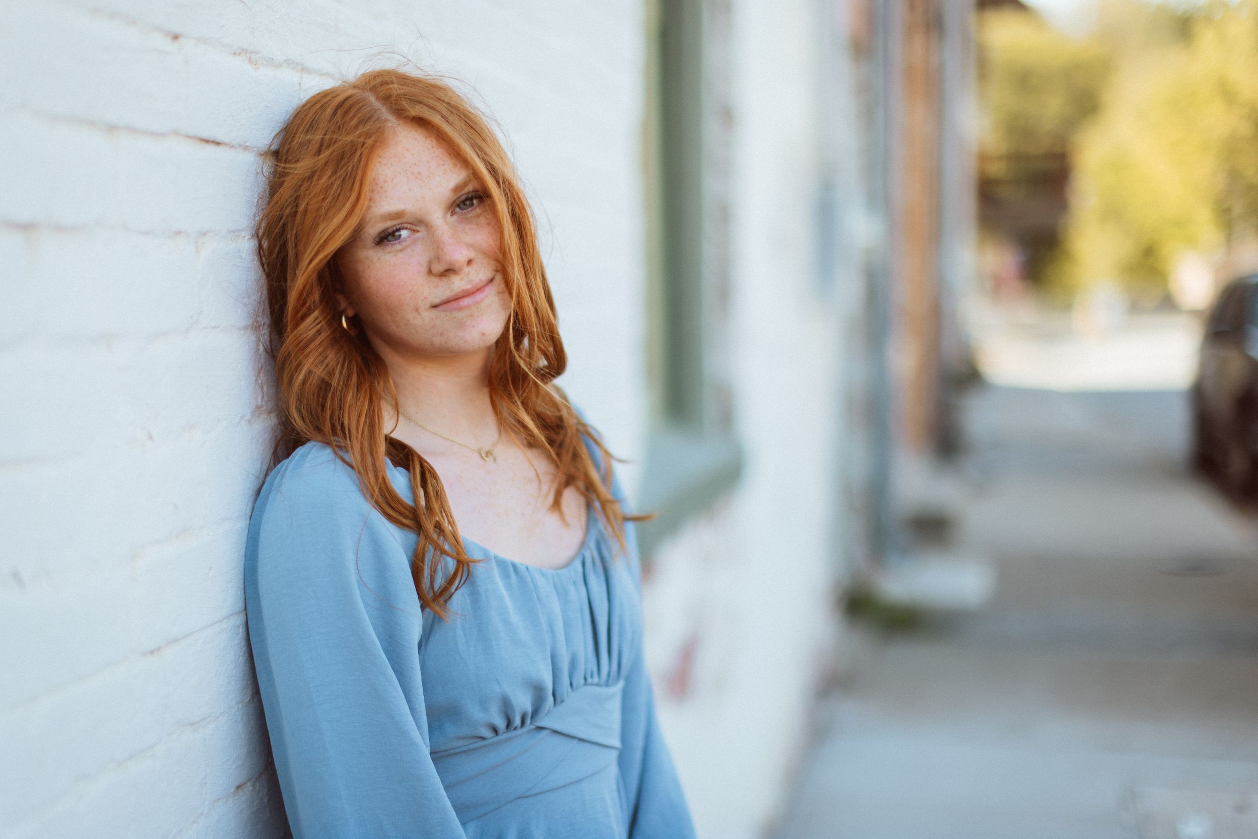 A red-headed woman in light blue mini dress leans against a white brick building and smiles softly at the camera.