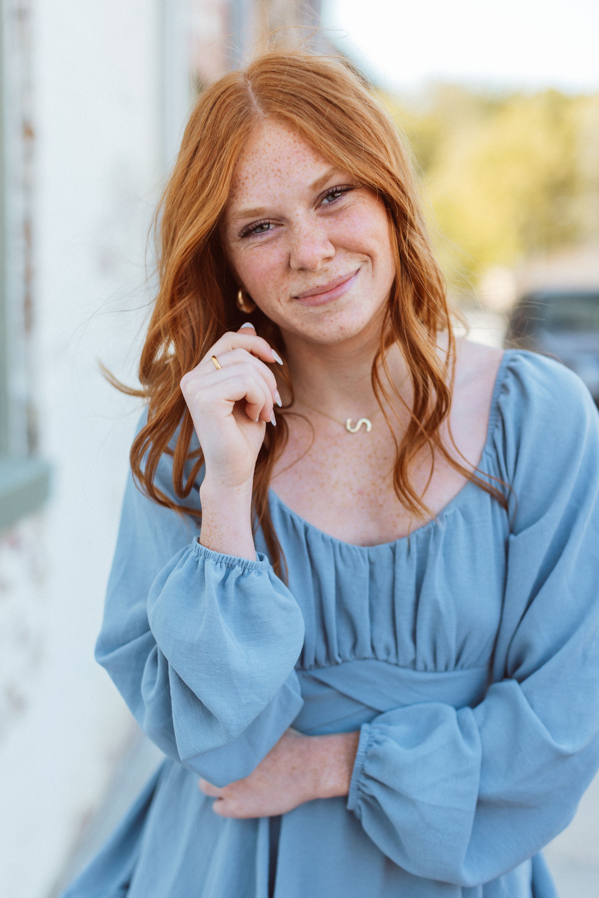 A red-headed woman in light blue mini dress stands in front of a white brick building and smiles softly at the camera.