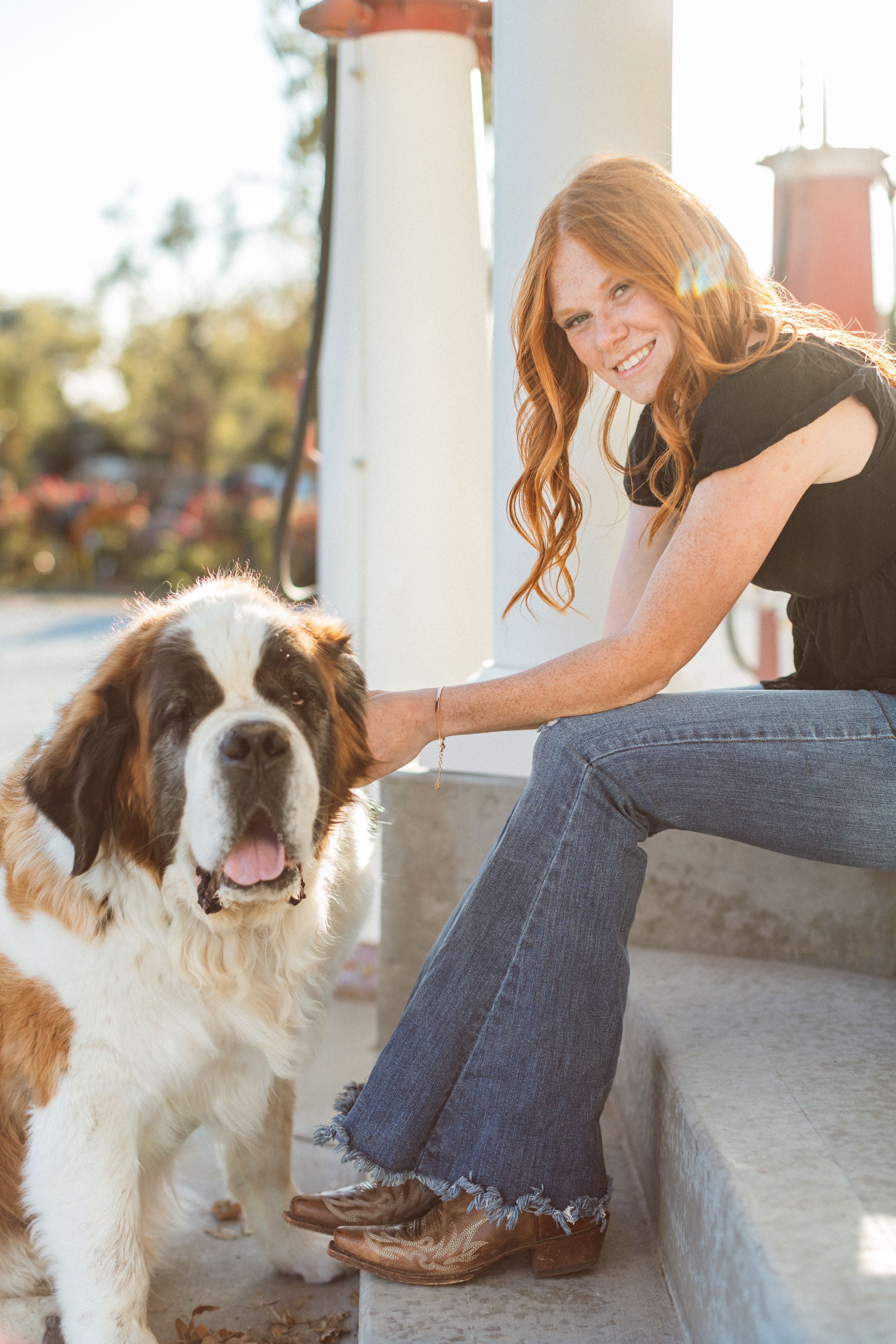 A red-headed woman in black top and flare jeans sits on concrete steps by an old gas pump and smiles at the camera and pets a Saint Bernard