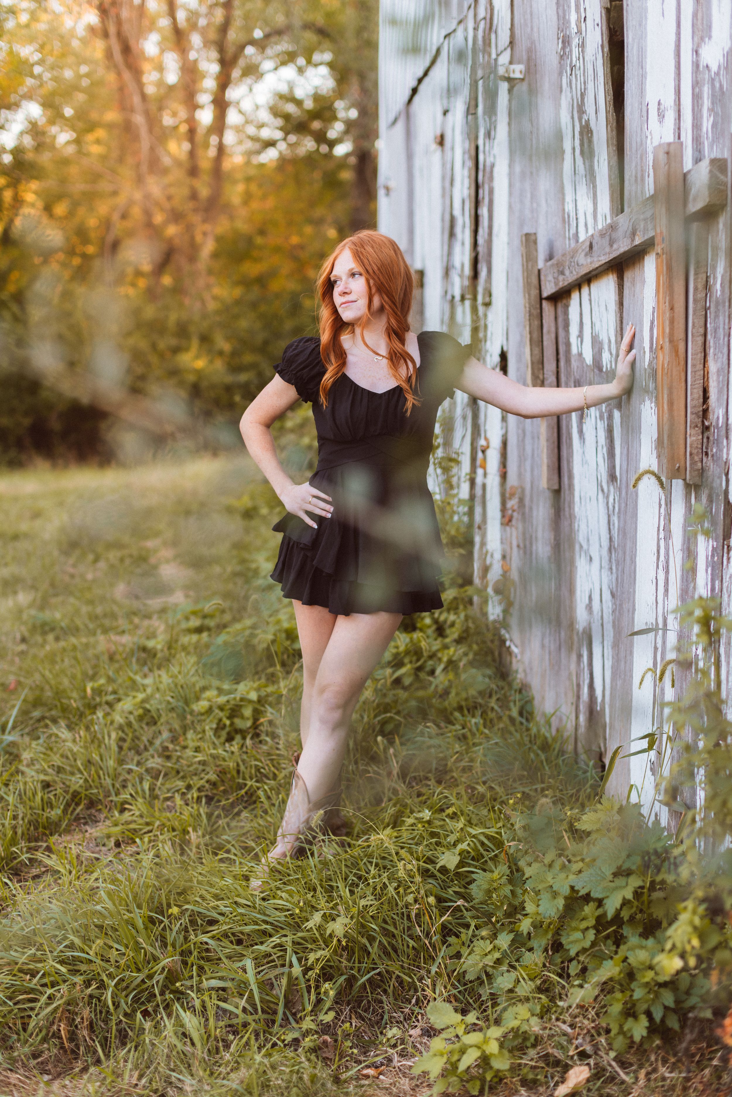 A redheaded girl wearing a black minidress leans against a rustic tobacco barn and looks away during her senior photo session in Kansas City