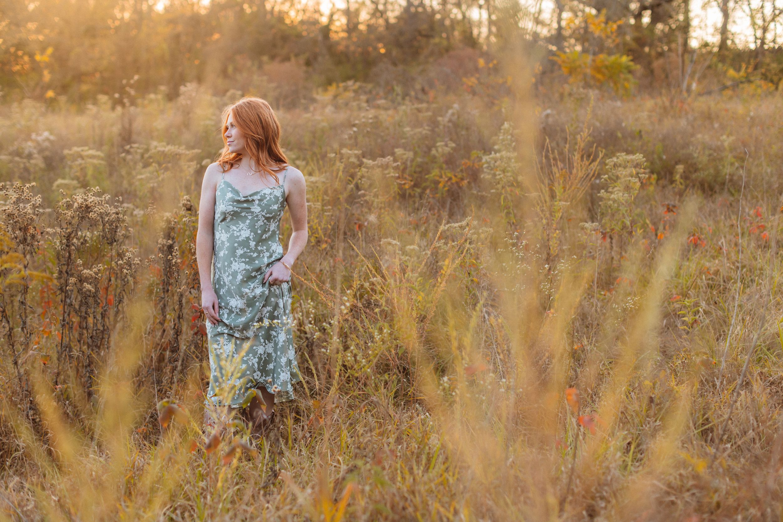 A red-headed young woman wearing a green floral dress stands in a golden field at sunset and looks over her shoulder into the distance.