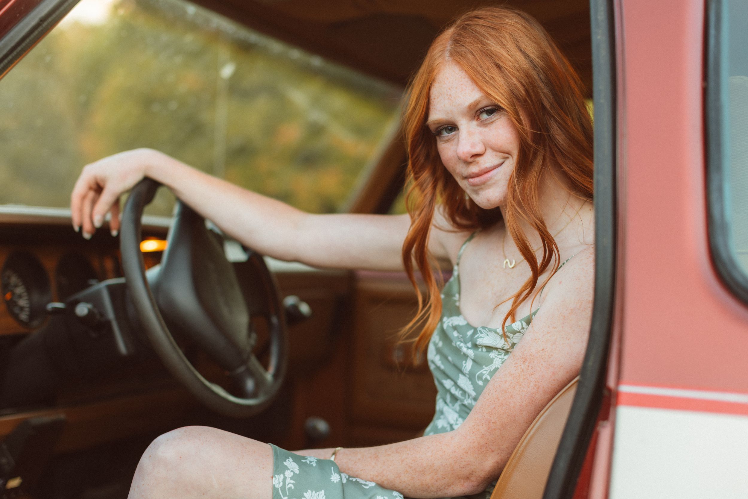 A red-headed young woman sits in an old pickup truck and rests her wrist on the steering wheel as she smiles softly at the camera.