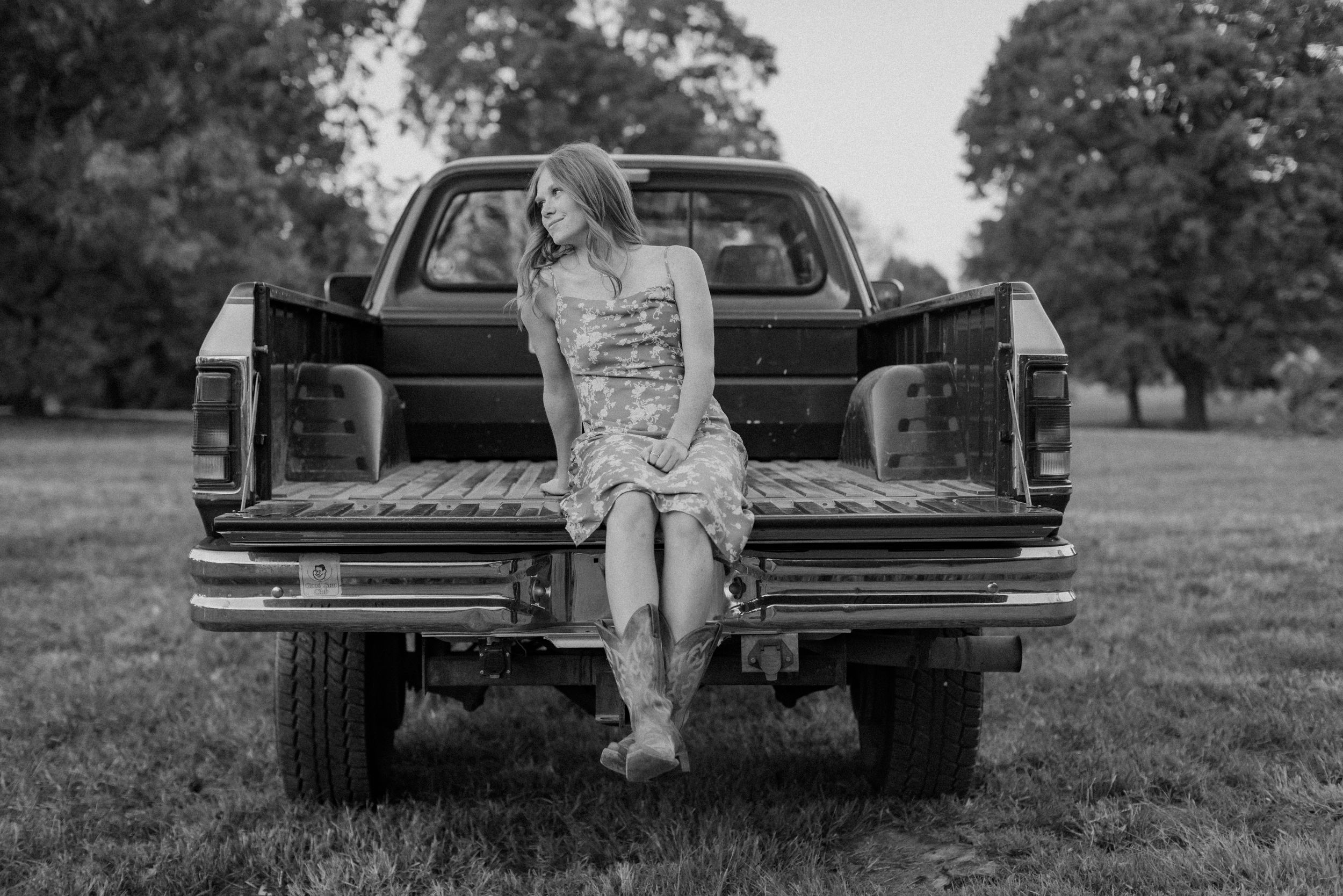 A young woman wearing cowboy boots and a sundress sits on the bed of a truck and looks off to the side during her Kansas City senior photos.
