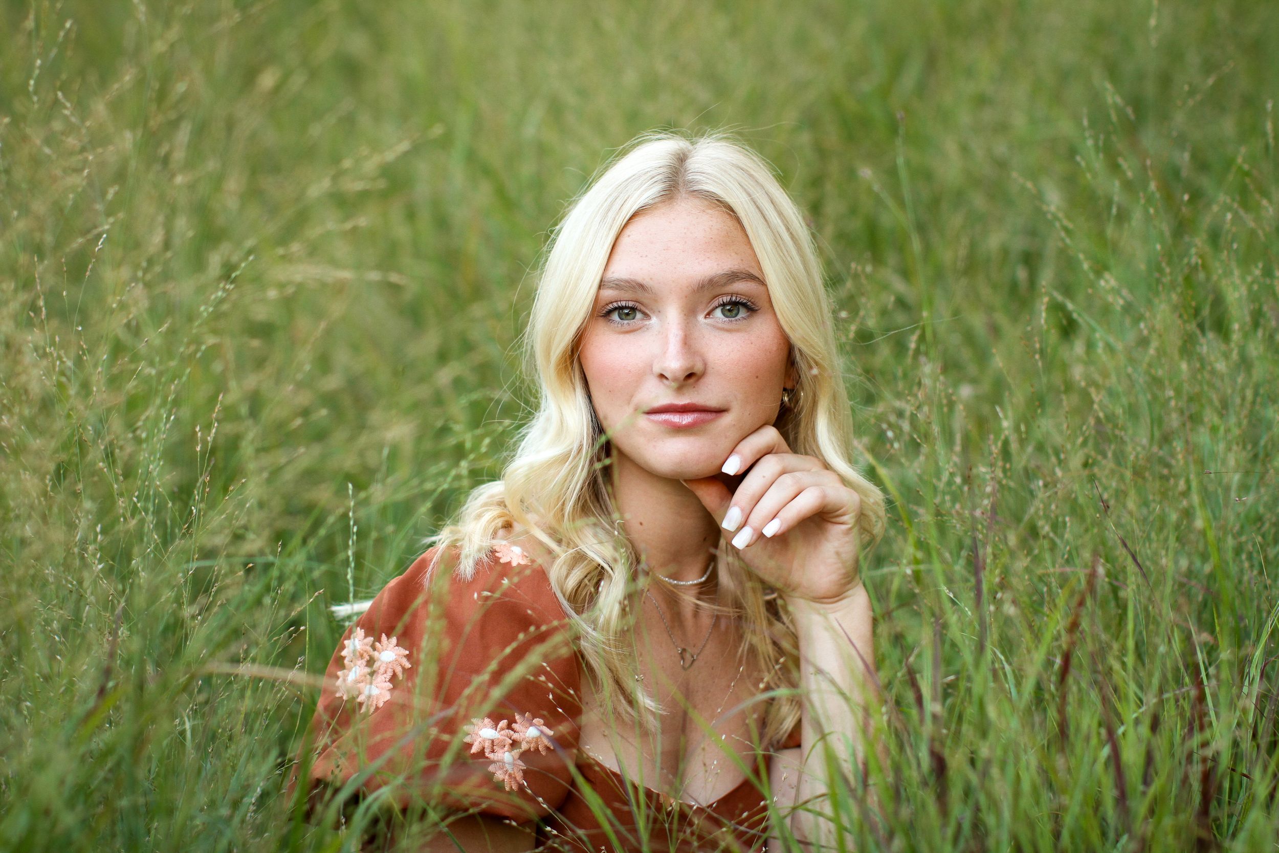 A young woman with blonde hair and green eyes rests her chin lightly in her hands in a tall, green grassy field during her KC senior photos.