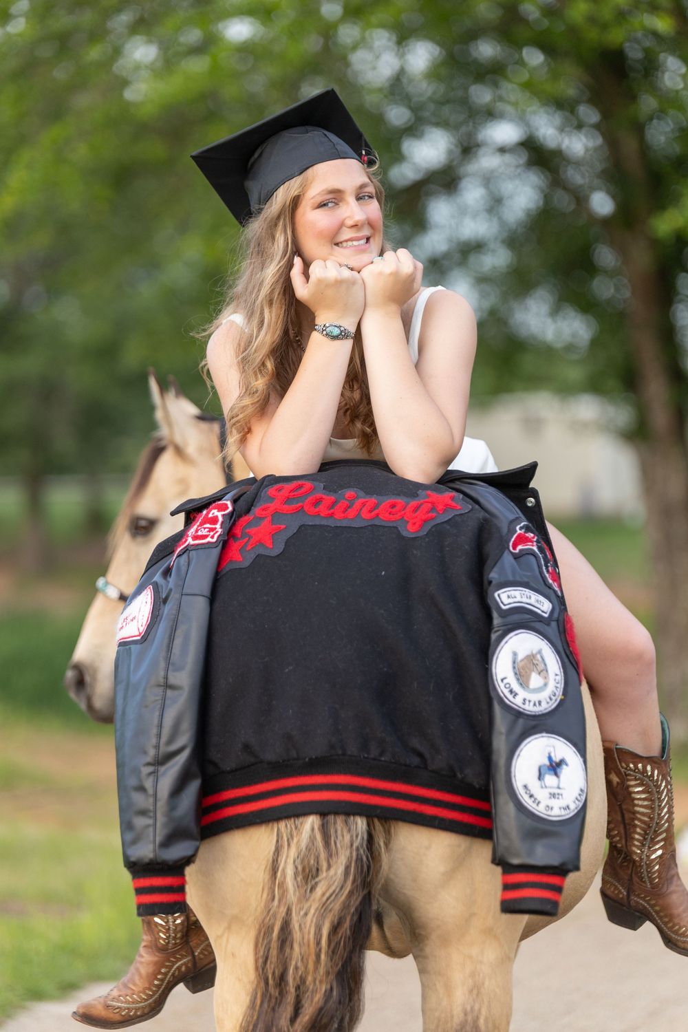 Girl sitting on her horse wearing her graduation cap, letterman jacket draped across the horse's rump, horse looking over it's shoulder