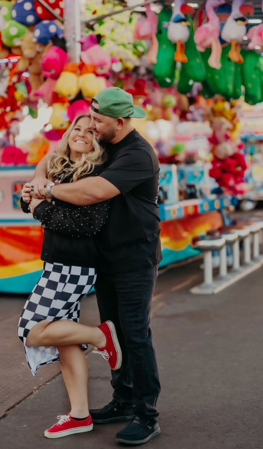 Couple embracing and smiling at a colorful carnival midway with stuffed animal prizes hanging in the background.