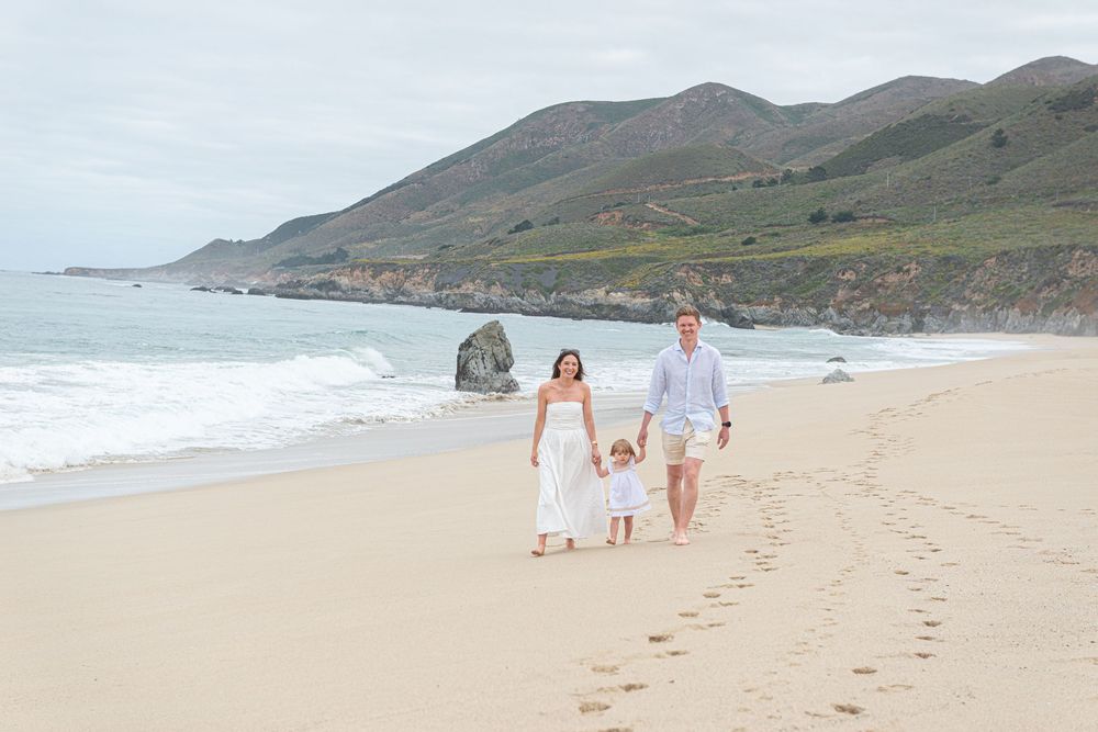 Family of three walking hand-in-hand on a scenic California beach with green hills and ocean waves in the background.