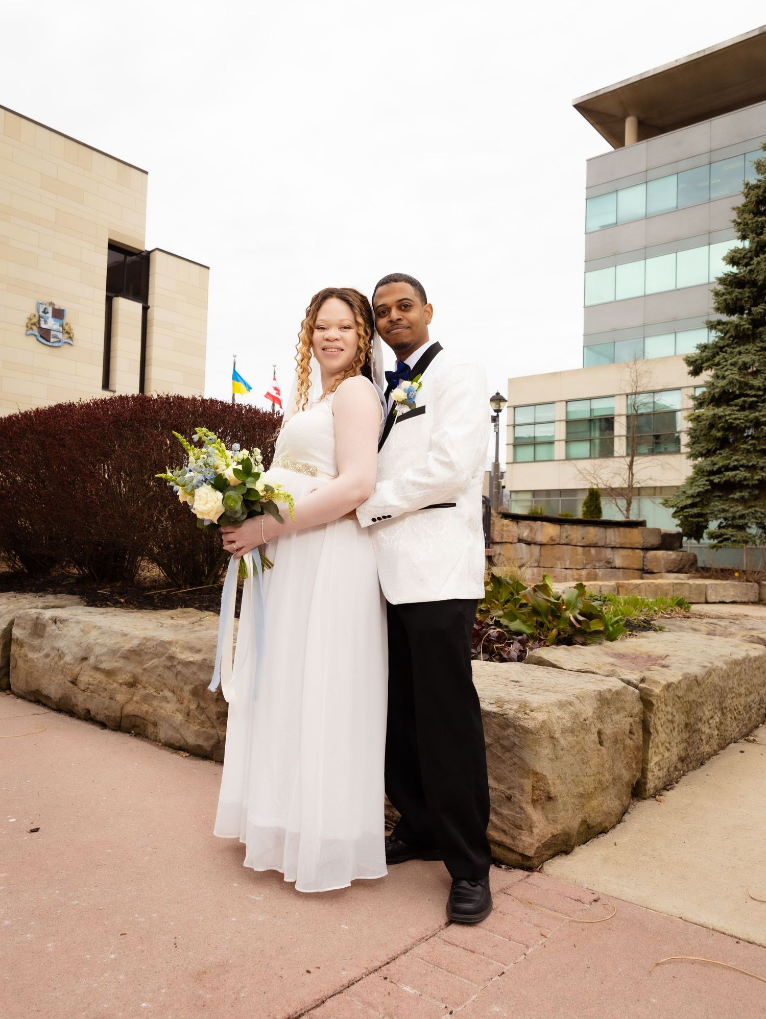 Newlywed couple posing outdoors in wedding attire, bride in white gown holding bouquet, groom in white tuxedo jacket.