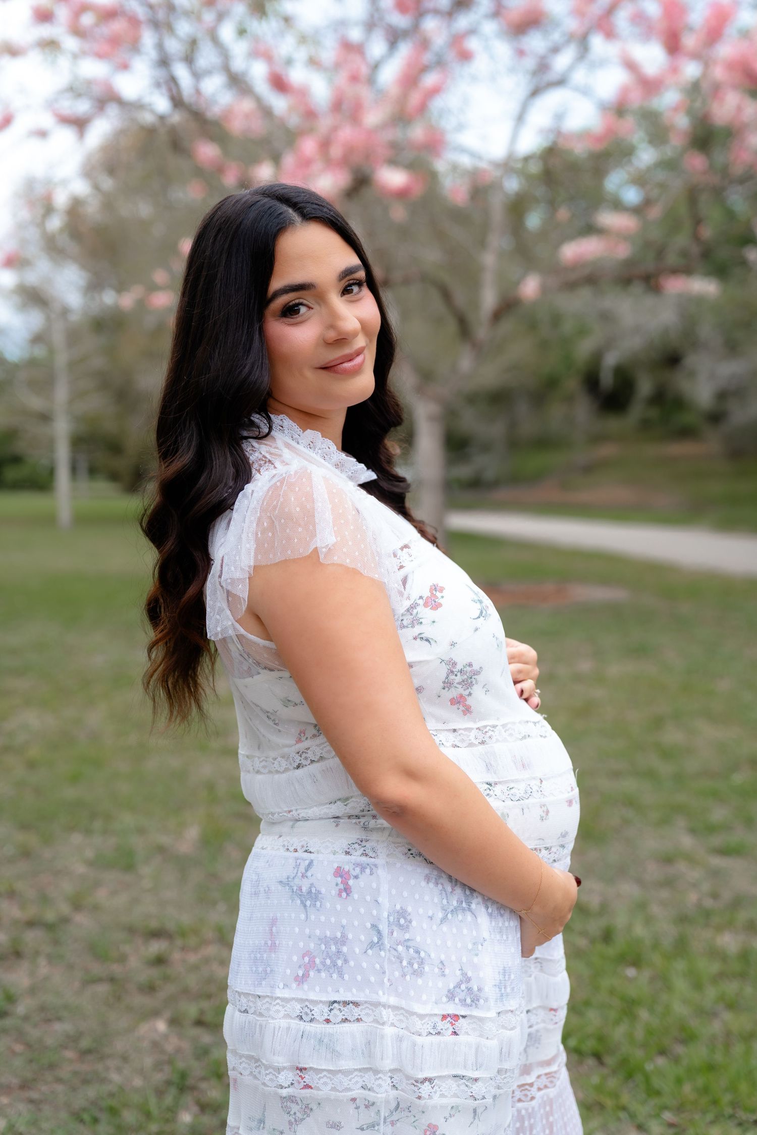 Pregnant woman in white lace floral dress cradling baby bump outdoors near blooming pink magnolia trees.