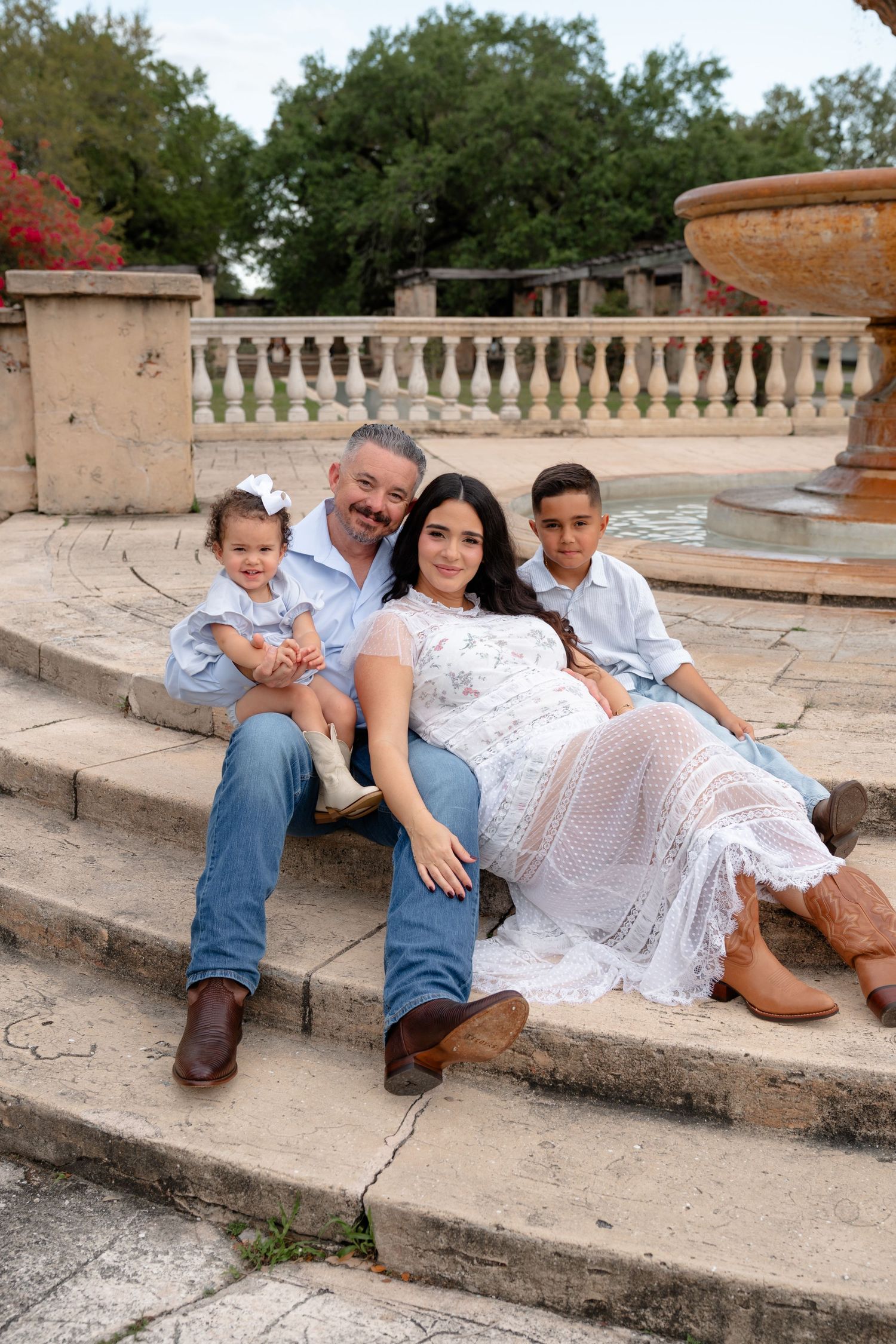 Family of four poses on stone steps near a fountain, mom in white lace dress, dad and kids in blue and jeans.