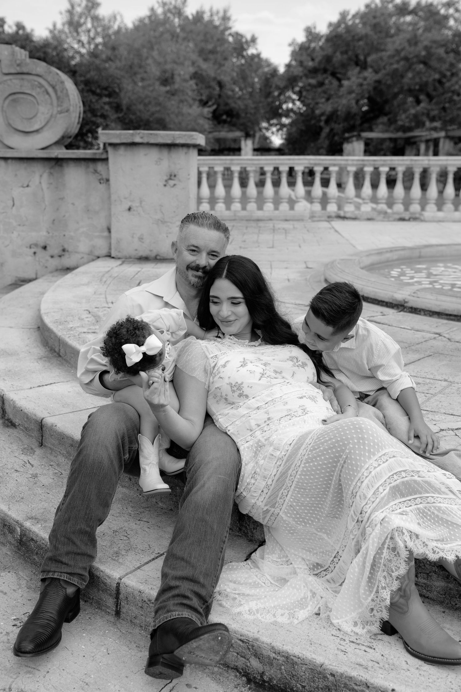 Black and white family maternity photo on stone steps, parents with two young children, elegant lace dress, outdoor setting.