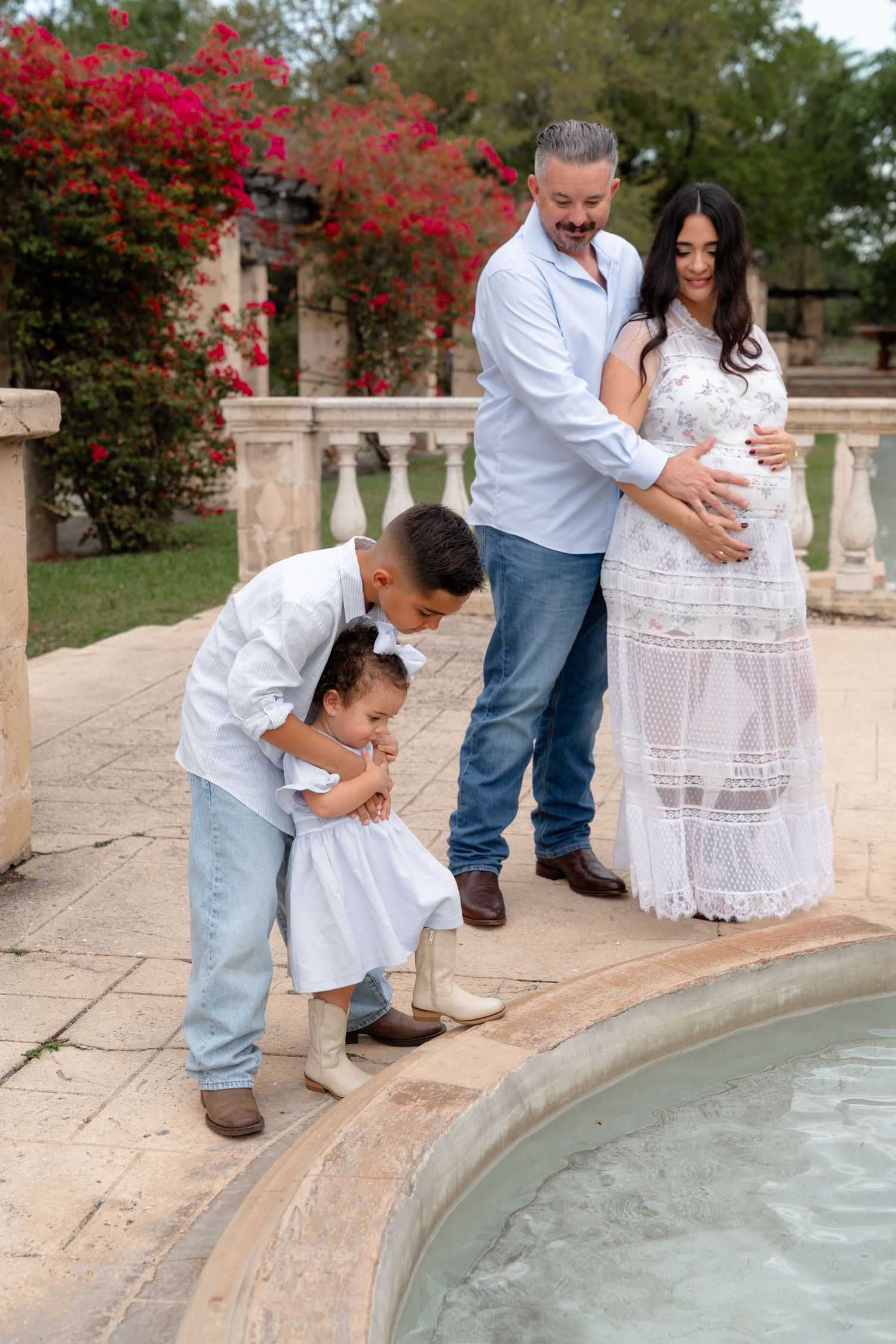 Pregnant woman in white lace dress with husband and two children by a fountain in an elegant outdoor setting.