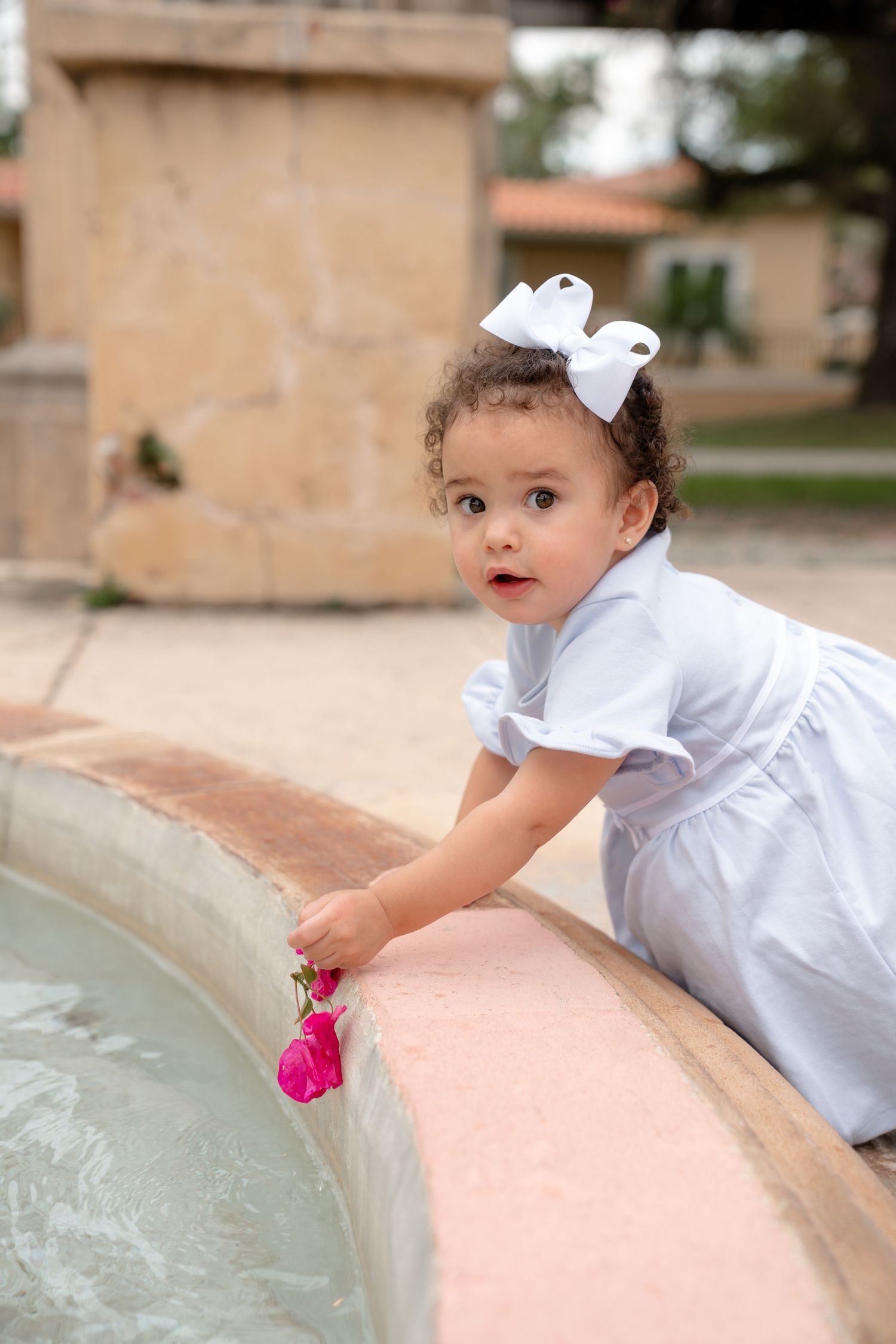 Toddler girl in white dress with bow holds pink flower at stone fountain outdoors.