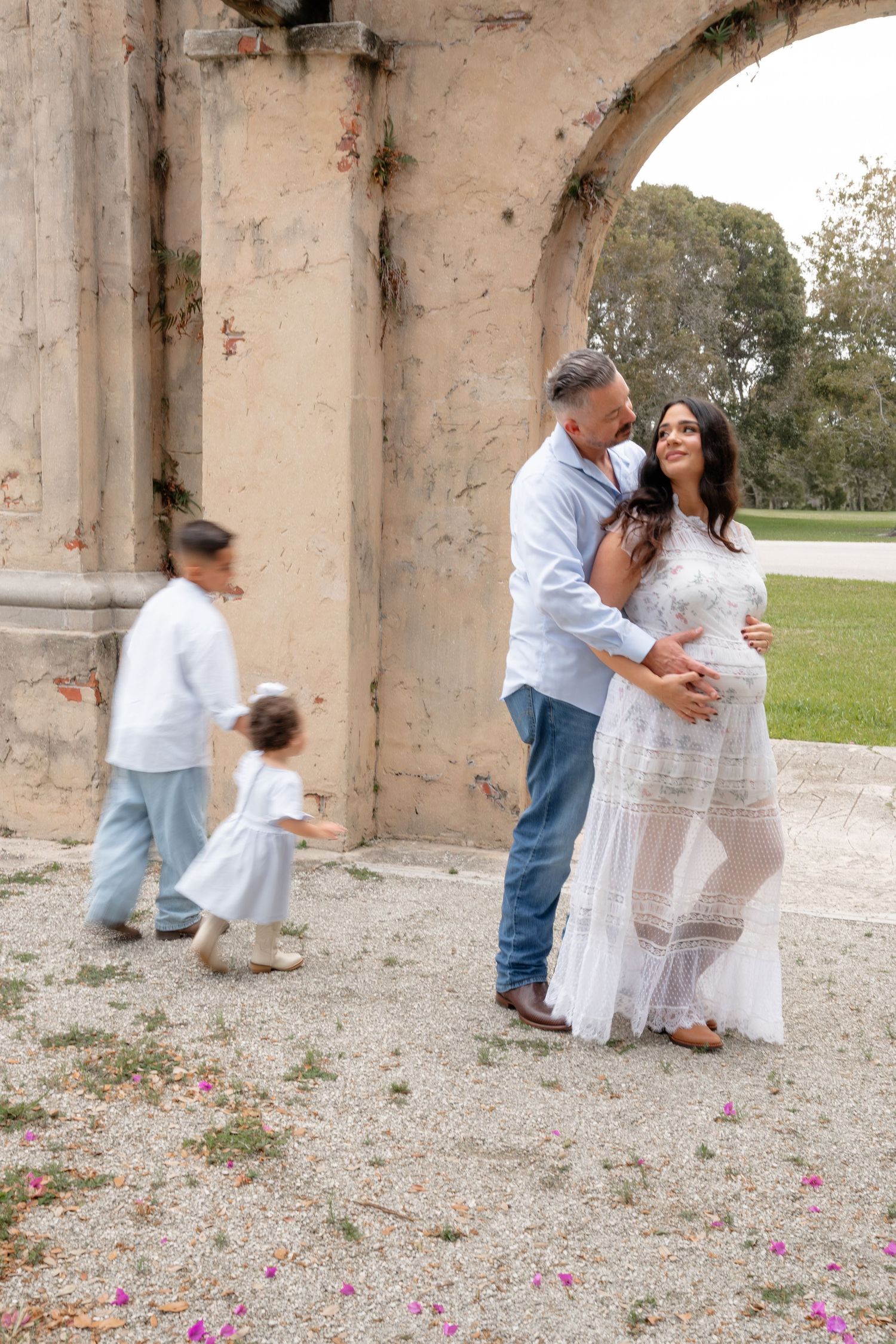 Pregnant woman in white lace dress with husband and children posing near stone arch outdoors.
