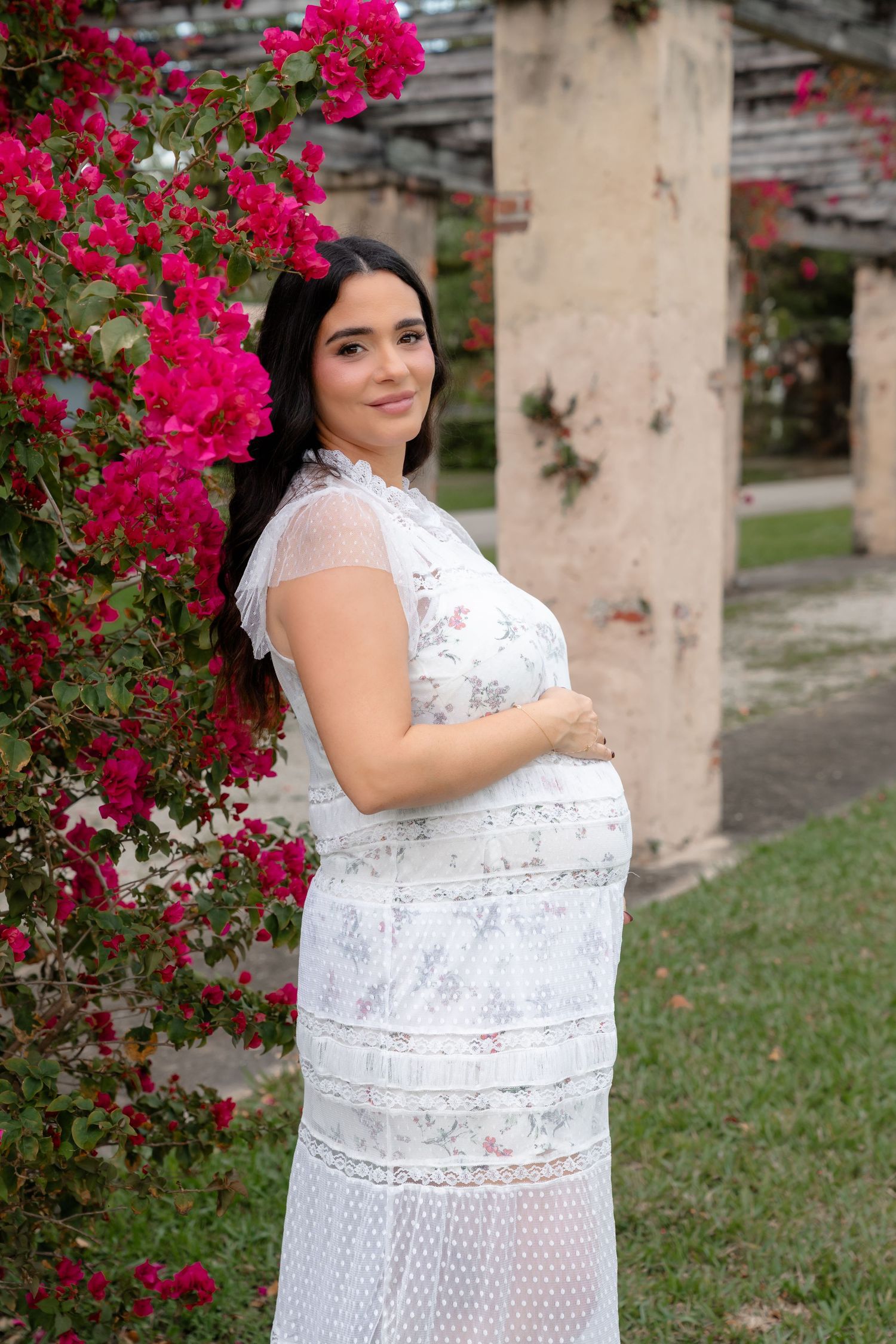 Pregnant woman in white lace floral dress posing beside vibrant pink bougainvillea flowers in a garden.