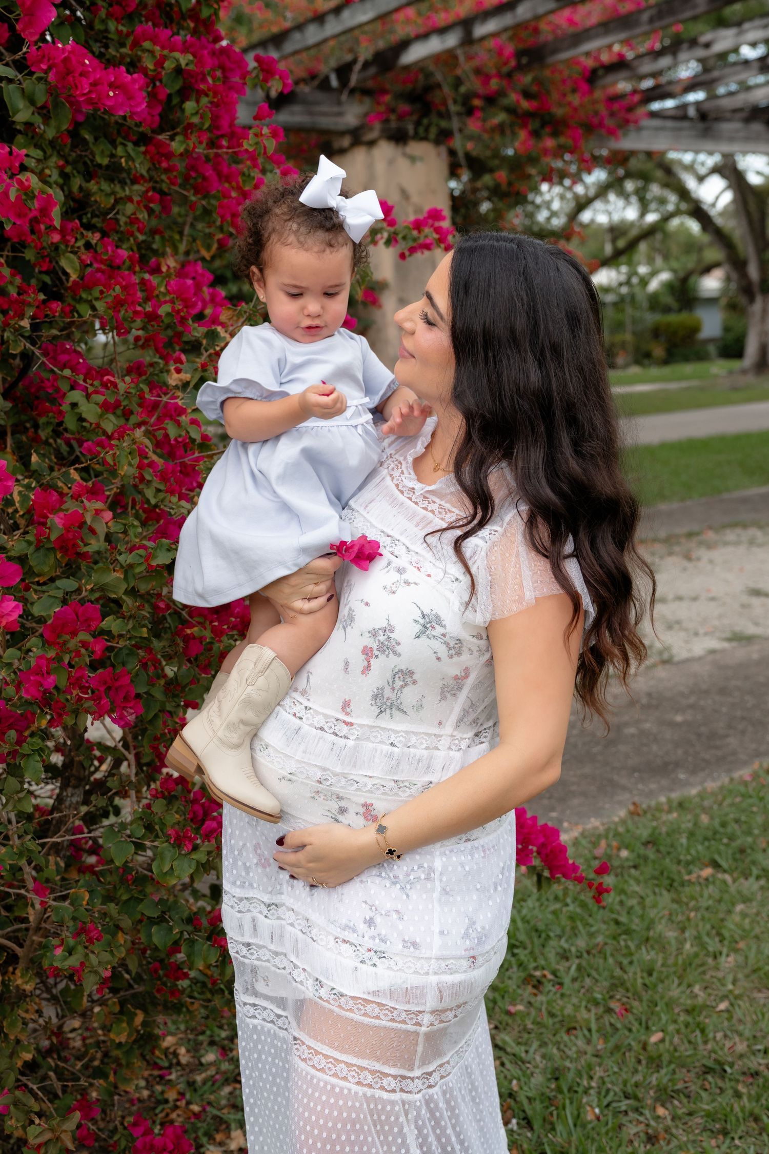 Pregnant woman in white lace dress holds toddler girl in blue dress near pink bougainvillea flowers outdoors.