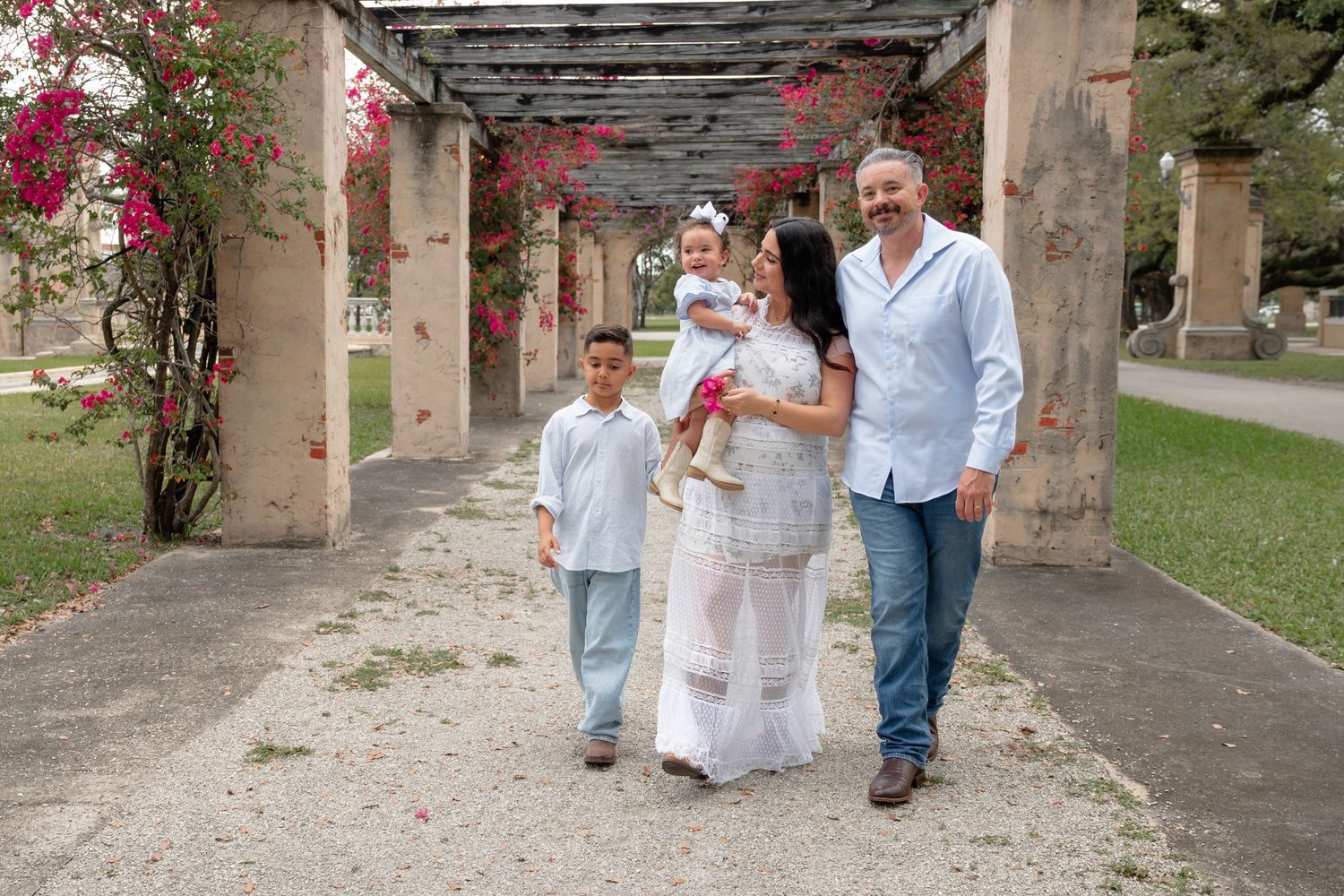 Family of four walking under flower-covered pergola in garden, parents with young children in coordinating white outfits.