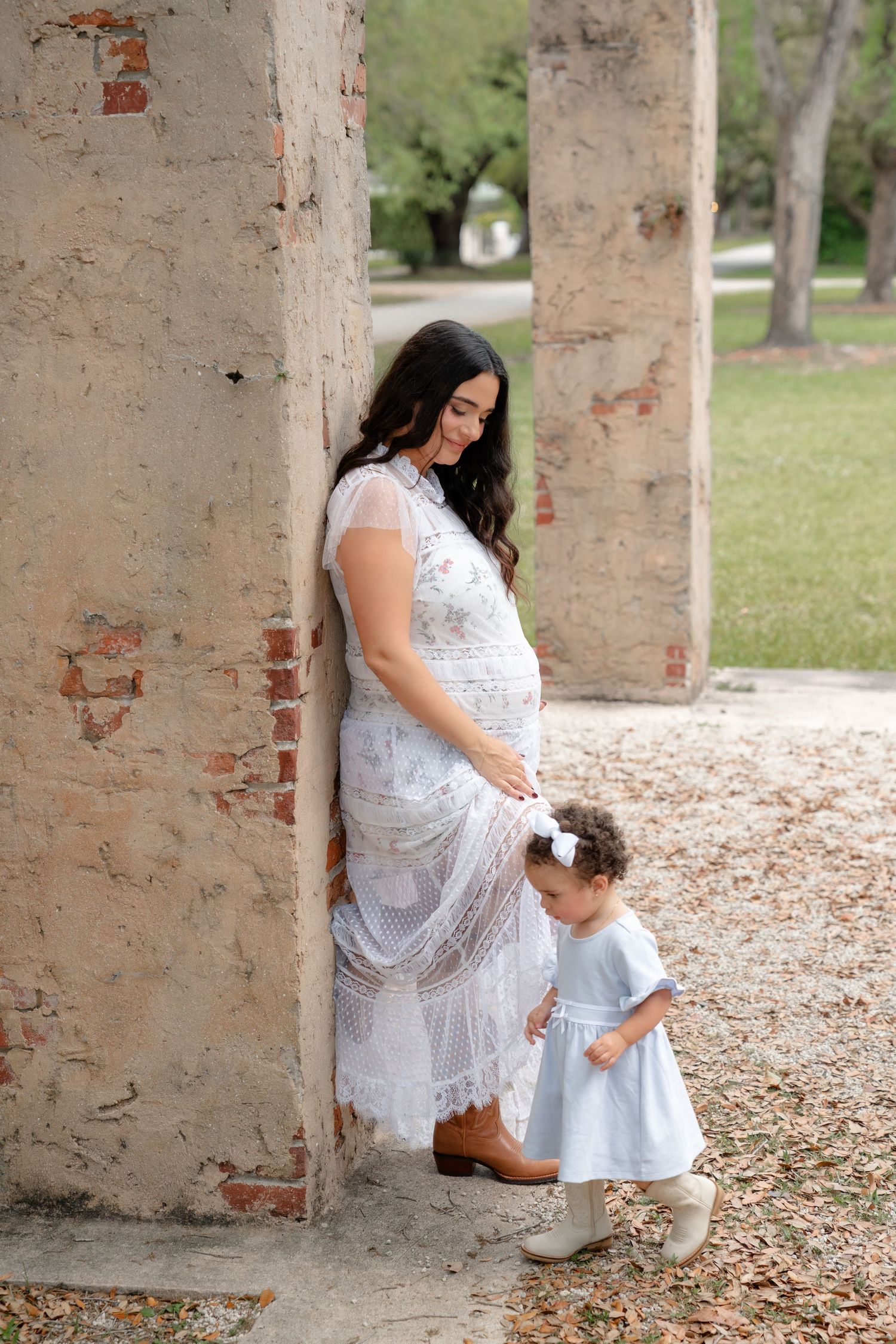 Pregnant woman in white lace dress with toddler girl in blue dress leaning against rustic brick column outdoors.