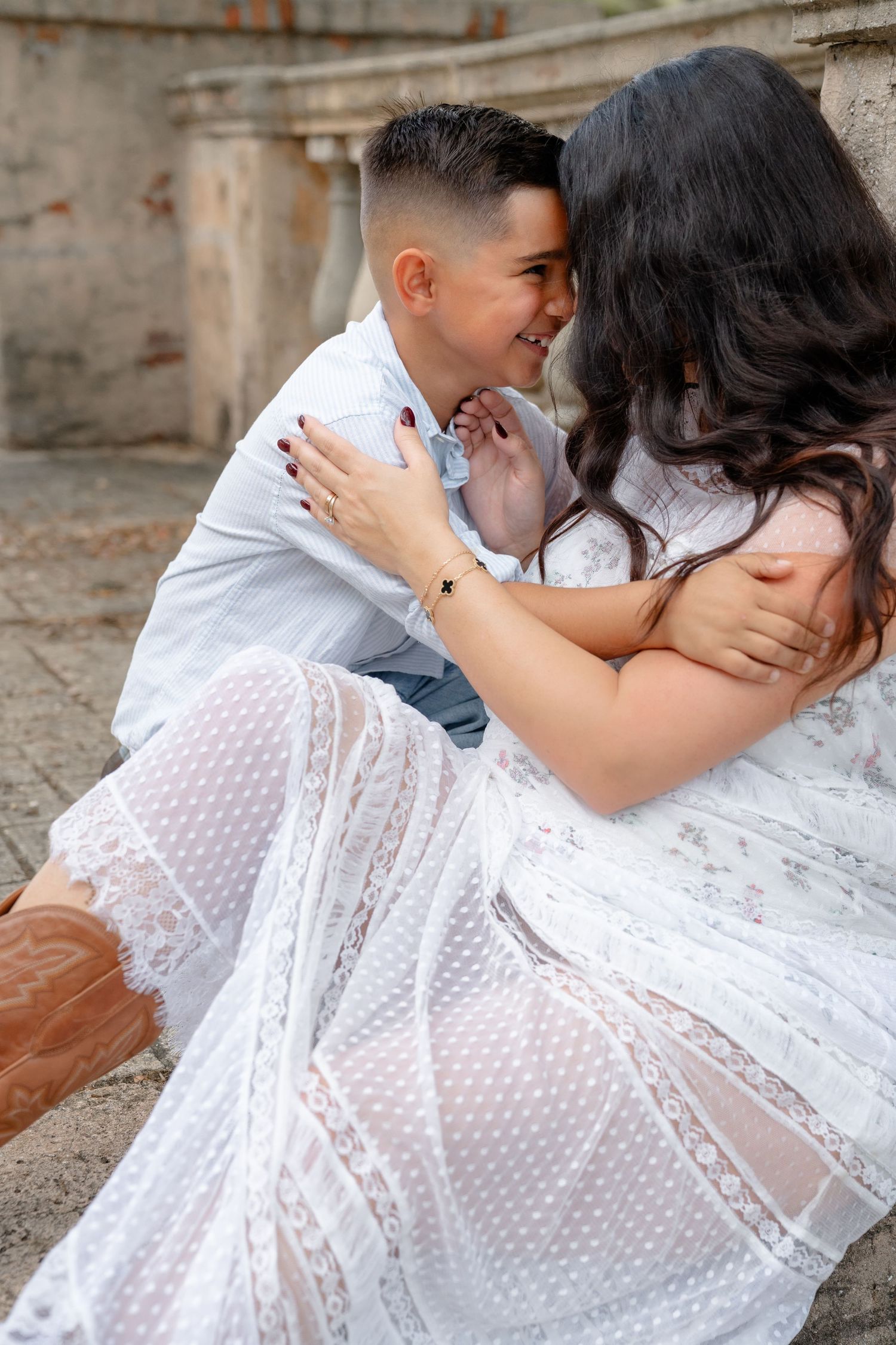 A mother in a white lace dress shares a tender moment with her young son on stone steps.