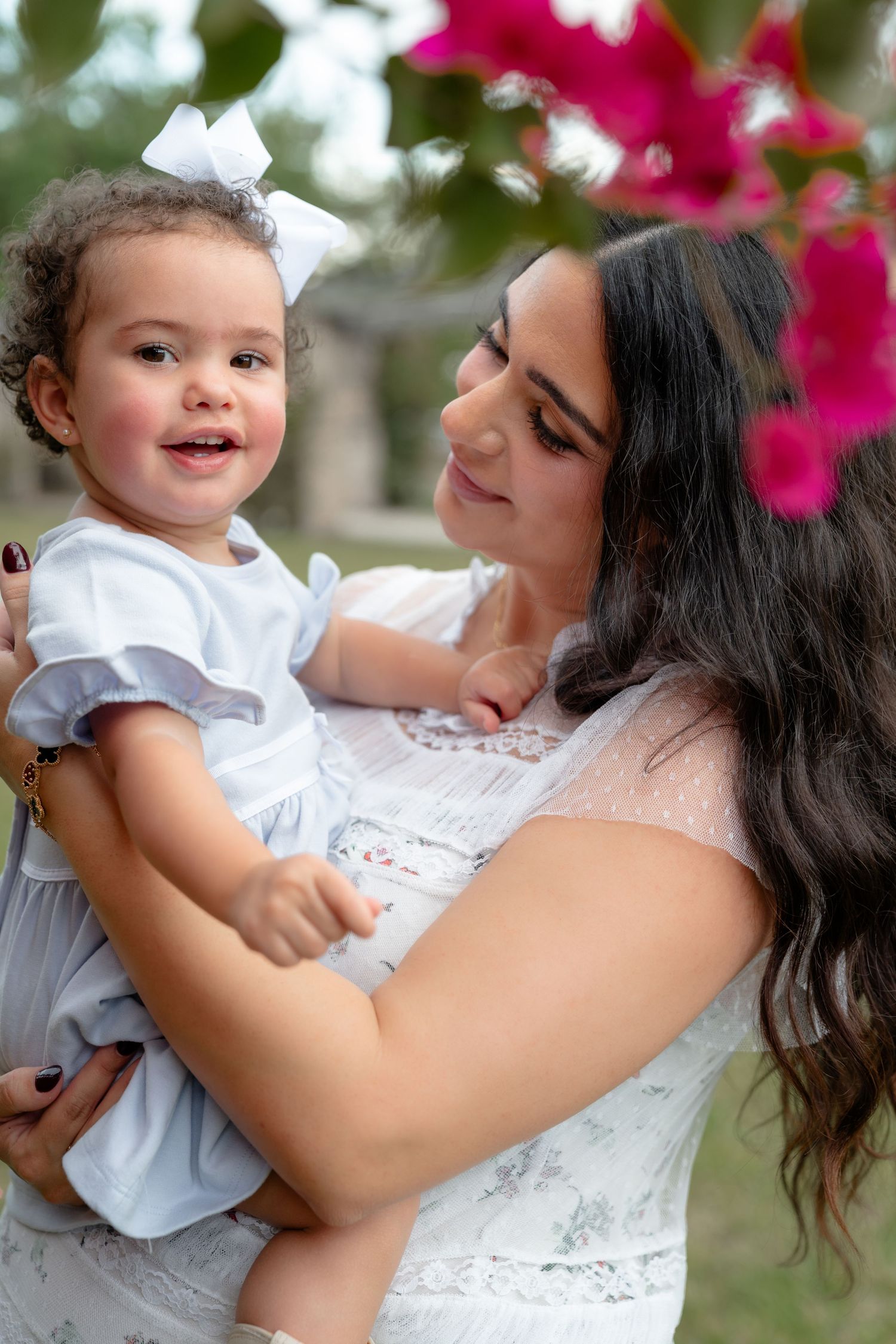 A young mother with dark hair holds her smiling toddler daughter in a white dress with a bow outdoors.