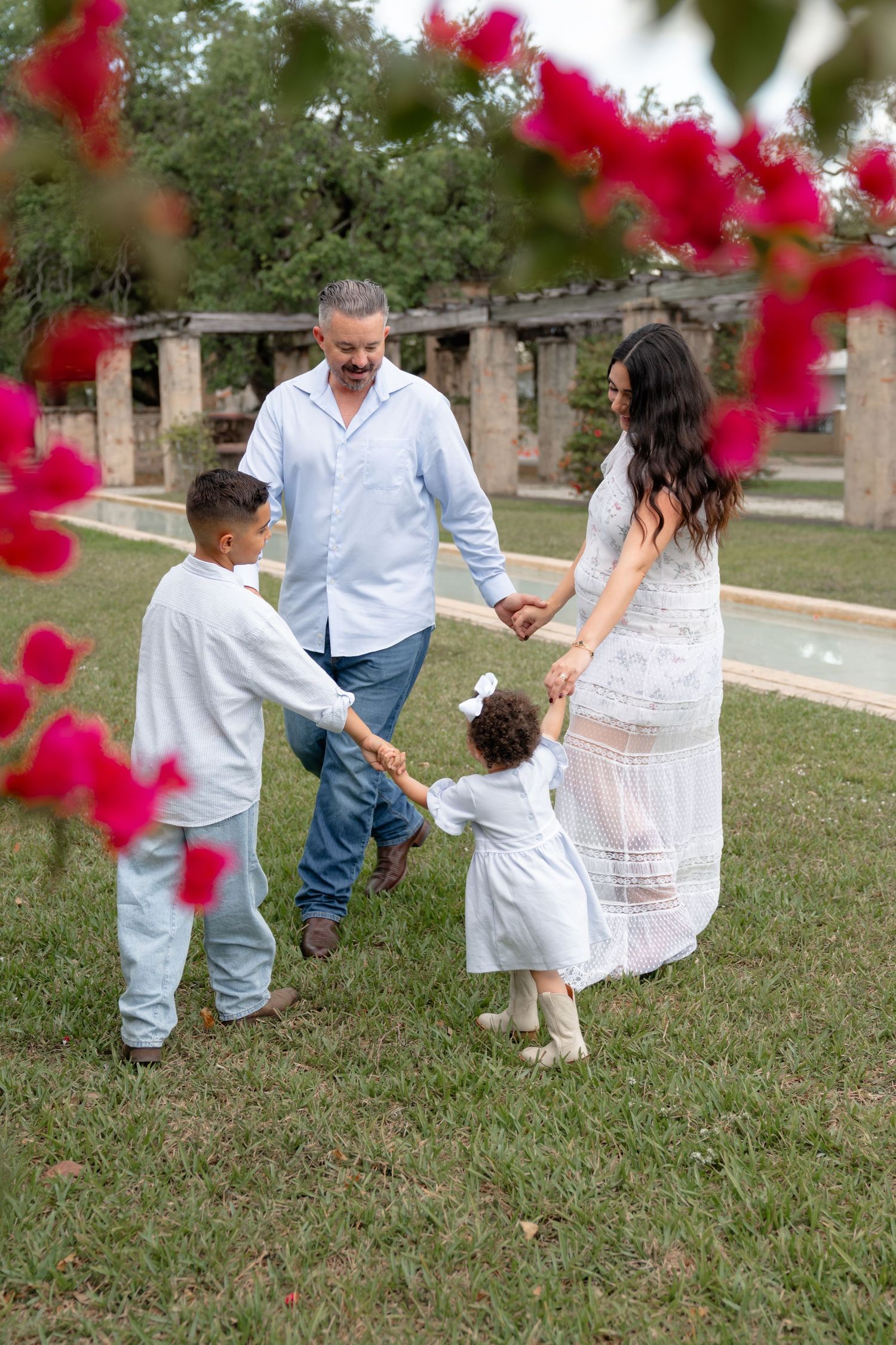 Family of four holding hands in a lush garden, framed by vibrant pink flowers, wearing white and blue outfits.