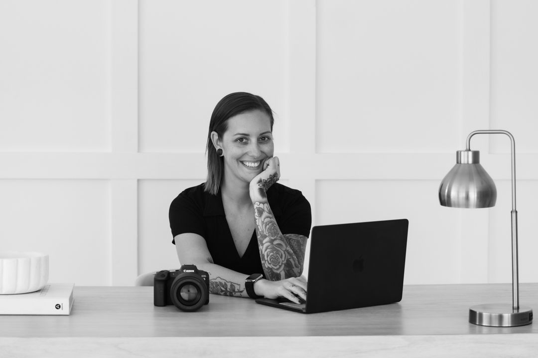 Black and white photo of a smiling woman with tattoos holding a camera, wearing a black top.