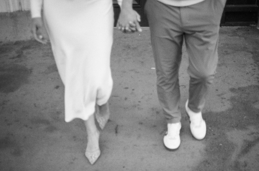 Black and white photo of a couple holding hands, woman in white dress and man in gray pants with white sneakers. 35mm film photoshoot.