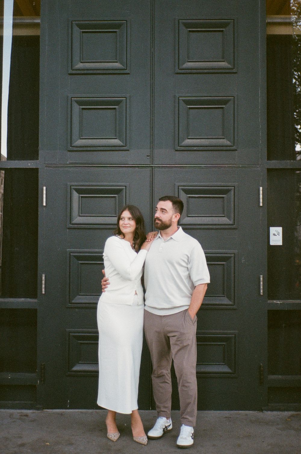 A couple poses elegantly in front of large black doors, woman in white dress, man in grey polo and tan pants. 35mm film engagement photos.