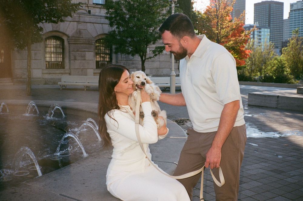 Couple sharing a tender moment with a small white dog near city fountains on a sunny autumn day. Engagement photoshoot Aliza Dworkind.