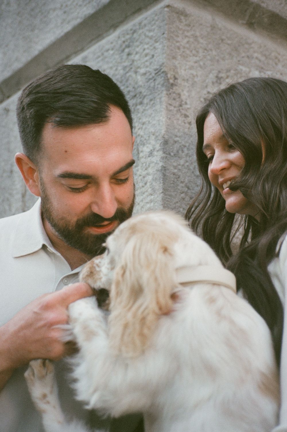 A couple smiling and bonding with a fluffy white Cocker Spaniel puppy against a stone wall backdrop. Captured on 35mm film during engagement