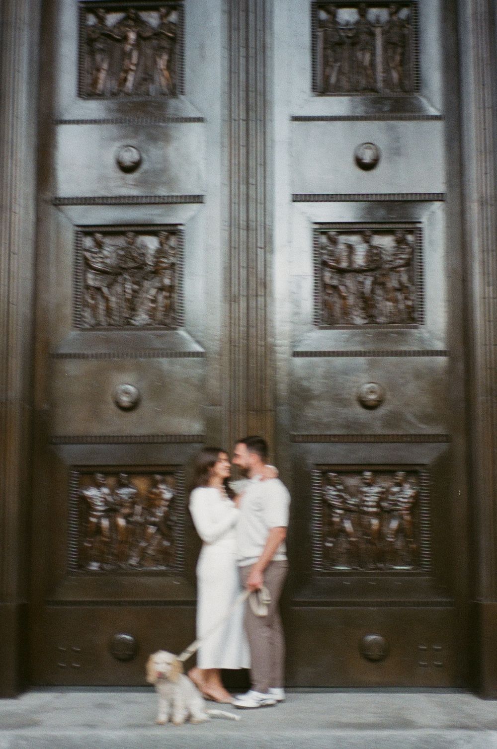 Couple embracing with a small dog before ornate bronze door. Captured on 35mm film during engagement photoshoot by Aliza Dworkind.