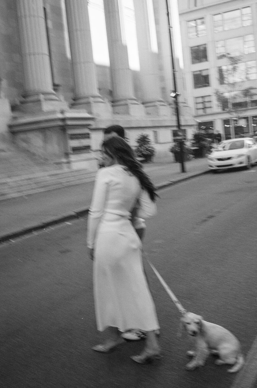 Black and white photo of a woman in a white dress walking a small dog on a leash past grand stone columns. Captured on 35mm film photoshoot.