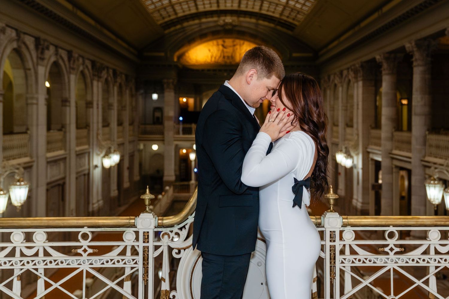 Couple sharing a romantic moment in an elegant grand hall with ornate architecture and warm golden lighting.