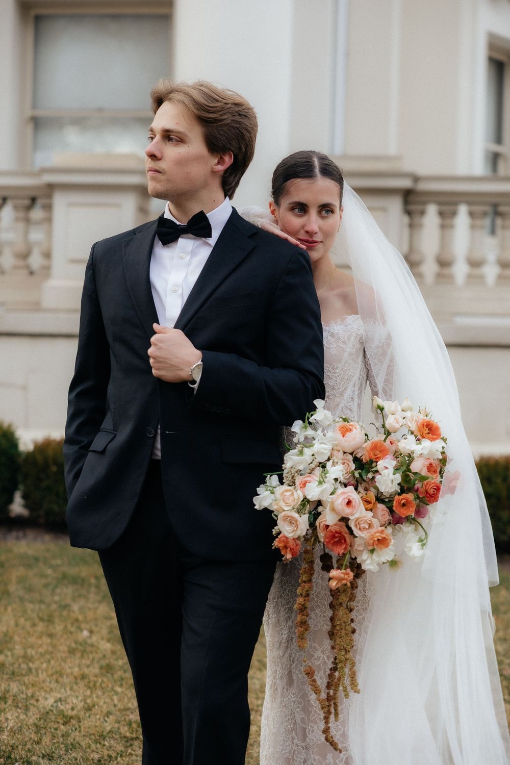 Bride and groom pose formally outside elegant building, she holds a cascading peach bouquet.