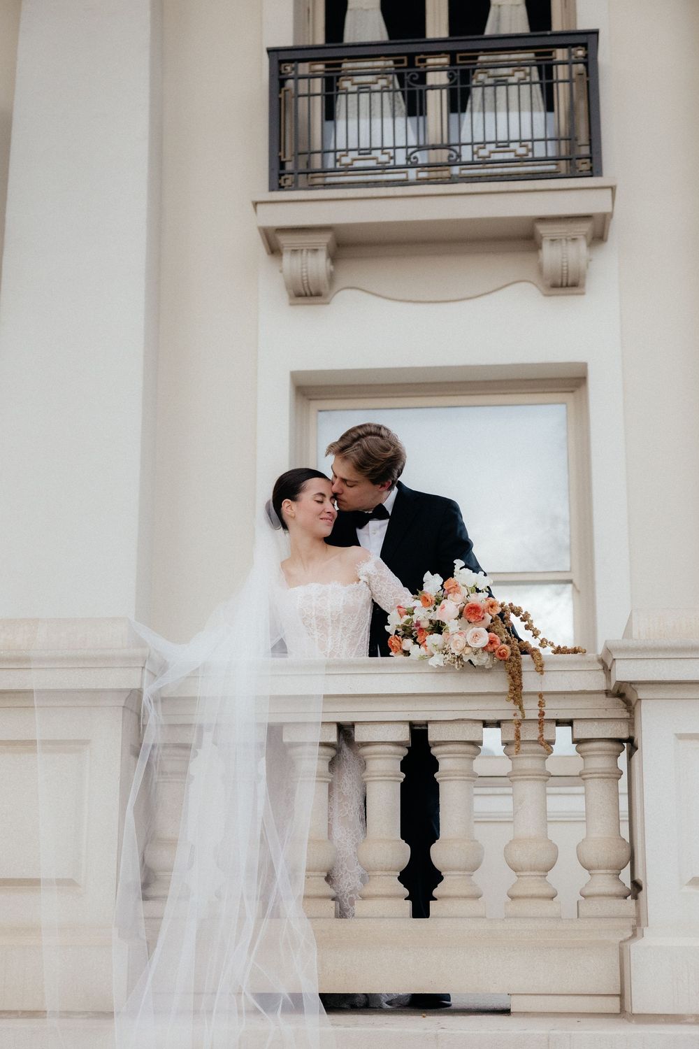 Bride and groom sharing an intimate moment on ornate balcony, bride in lace gown with flowing veil and bouquet.