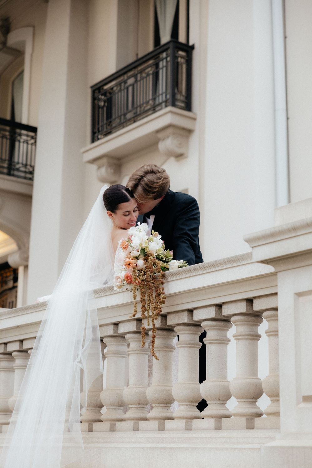 Bride in lace gown with flowing veil and groom in black suit share intimate moment on ornate white balcony.