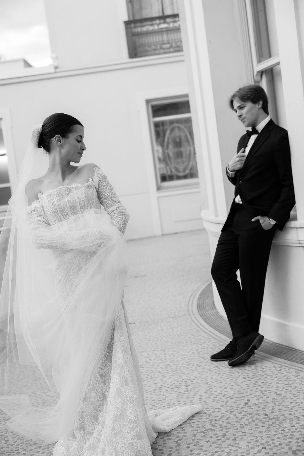 Black and white photo of bride in lace gown with veil and groom in tuxedo posing at elegant venue.