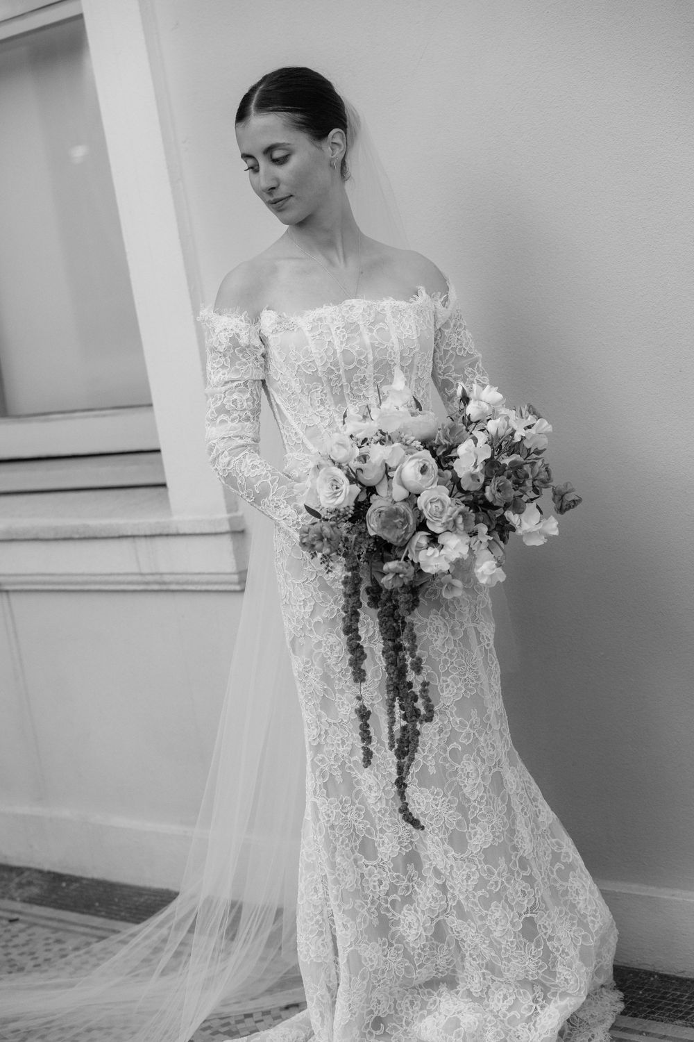 Black and white full-length portrait of bride in lace gown holding cascading floral bouquet by column.